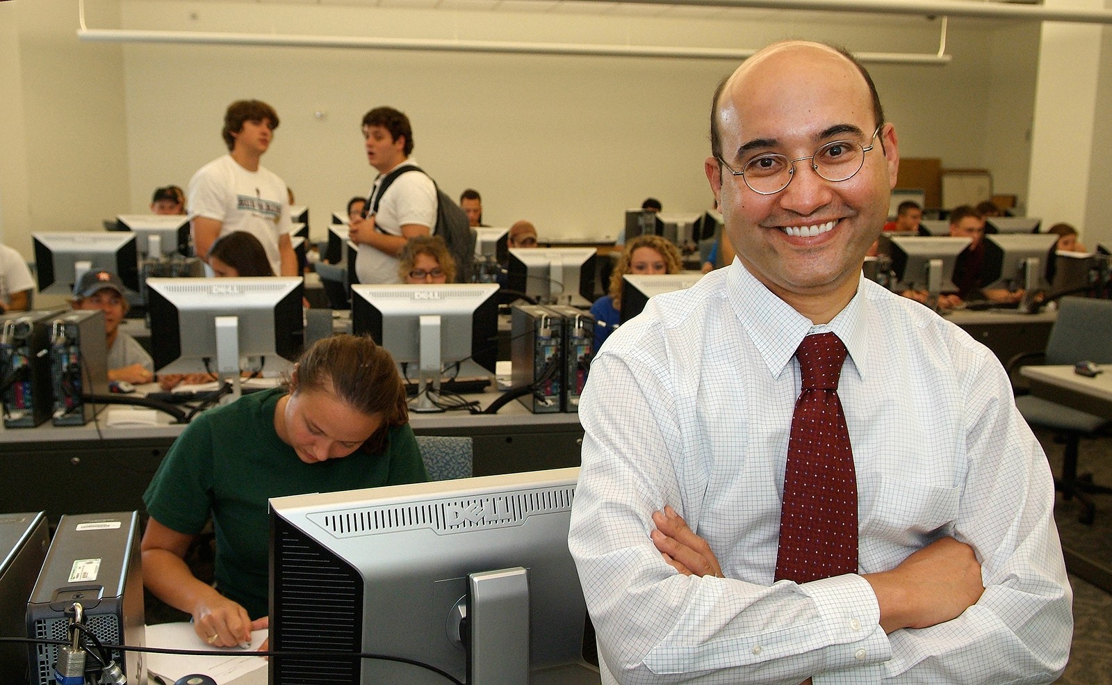 Dr. Mahmoud El-Halwagi stands with his arms crossed and smiles in front of a classroom with computers and students working and socializing. He's wearing a white button-down shirt and a red tie. 